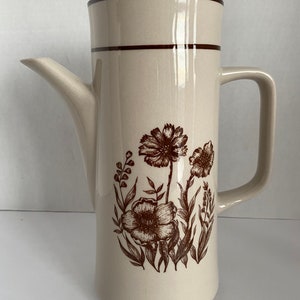 May include: A brown and white ceramic coffee pot with a floral design. The pot has a brown lid and a brown handle.