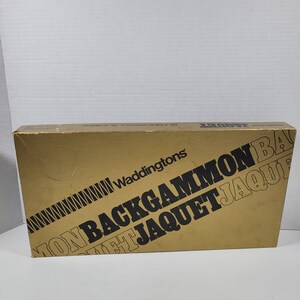 Può includere: Una scatola dorata con testo nero che dice "Waddingtons Backgammon Jaquet Jaquet" e "1981 A.R.B.G. (UK) Toys & Games Ltd."