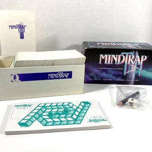Könnte beinhalten: Eine Mind Trap Brettspielbox mit einem weißen und blauen Design. Die Box ist geöffnet und zeigt einen Stapel Karten. Das Spiel beinhaltet ein Spielbrett, Würfel und einen Bleistift.