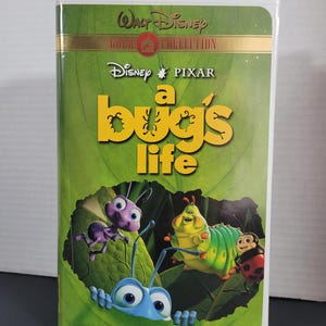Peut inclure: Une cassette VHS pour le film Disney Pixar "1001 pattes". Le boîtier est blanc avec un fond vert et présente un gros plan des personnages Flik, Heimlich et une coccinelle.