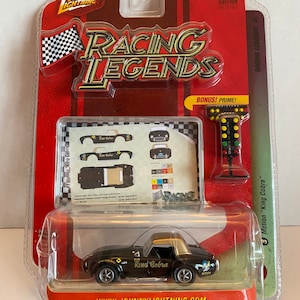 Peut inclure: Une miniature de voiture de course en métal moulé sous pression noire et dorée représentant une Shelby Cobra de 1965, faisant partie de la série Racing Legends de Johnny Lightning. La voiture a une carrosserie noire avec des accents dorés, un toit doré et les mots "King Cobra" imprimés sur le côté. La voiture est présentée sur une carte rouge et blanche avec les mots "Racing Legends" imprimés dessus.