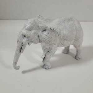 May include: A white ceramic elephant figurine with a textured surface. The elephant is standing with its trunk raised and its head tilted to the side.
