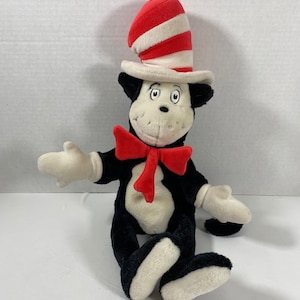 May include: A black and white plush cat wearing a red and white striped hat with a red bow tie. The cat is sitting with its legs crossed and its arms outstretched.