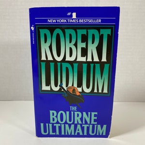 May include: A blue paperback book titled "The Bourne Ultimatum" by Robert Ludlum. The book is a New York Times bestseller and features a silhouette of the Capitol Building.
