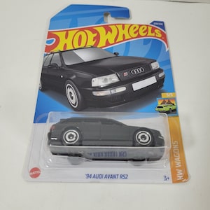 May include: A black 1994 Audi Avant RS2 Hot Wheels car in a plastic package. The car is a wagon with a black body and silver wheels. The package is blue and yellow with the Hot Wheels logo and the car's name and year.