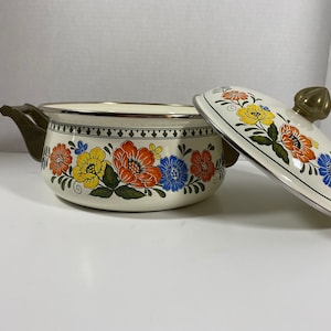 May include: A white enamel pot with a floral design in shades of orange, yellow, blue, and green. The pot has two brass handles and a matching lid with a brass knob.
