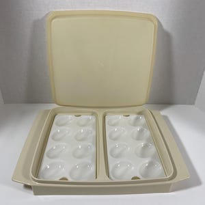 Puede incluir: Un porta huevos de plástico beige con tapa. El porta huevos tiene dos secciones, cada una con seis huecos en forma de huevo. La tapa es ligeramente abovedada.