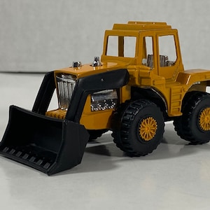 Puede incluir: Un tractor de juguete amarillo con una pala frontal negra. El tractor tiene una cabina negra con un parabrisas transparente y un asiento negro. El tractor tiene ruedas negras con llantas doradas.