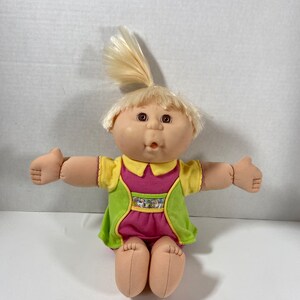 May include: A Cabbage Patch Kid doll with blonde hair, brown eyes, and a pink and green outfit. The doll is sitting with its arms outstretched.