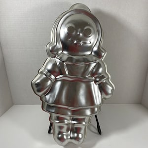 Op de afbeelding: Een zilveren metalen poppencakevorm. De vorm is gevormd als een pop die een jurk draagt en heeft een eenvoudig gezicht met ogen, een neus en een mond.