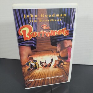 May include: VHS video cassette cover featuring John Goodman and Jim Broadbent in the film "The Borrowers". The cover has a blue and white striped background with the title in large, red letters. The tagline reads "Little People...Big Adventure."