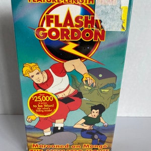 May include: A VHS tape case for the animated movie "Flash Gordon: Marooned on Mongo". The case features a colourful illustration of Flash Gordon, Dale Arden, and a green alien. The text "Over £25,000 in prizes to be won!" is on a yellow circle.