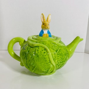 May include: A novelty green ceramic teapot in the shape of a cabbage, topped with a rabbit figurine. The rabbit is dressed in a blue jacket and yellow waistcoat. The teapot has a curved handle and spout.