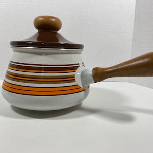 Peut inclure: Une casserole en émail blanc avec un couvercle brun et une poignée en bois. La casserole est ornée de rayures orange et brunes autour du corps.