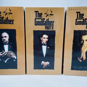 以下が含まれることがあります： ゴッドファーザーの映画3本のVHSテープ。ゴッドファーザー、ゴッドファーザーパートII、ゴッドファーザーパートIII。テープは金色で、黒い文字と主要キャラクターの画像が描かれている。