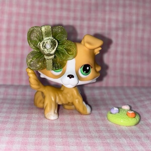 May include: A tan and white toy dog figurine with a green bow on its head and green eyes. The dog is kneeling next to a small green base with colorful, round objects on top. The background is a pink and white checkered pattern.