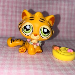Könnte beinhalten: Gelbe Tigerfigur mit schwarzen Streifen, großen grünen Augen und einer Blumenkette. Der Spielzeugtiger sitzt neben einem gelben, gewirbelten Objekt mit rosa und orangefarbenen Akzenten. Der Hintergrund ist ein rosa gemusterter Stoff.