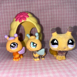 May include: Three yellow and orange bee-themed figurines, including a beehive house, a bee with wings, and a bee with a fuzzy body. The beehive house has a pink flower on top.