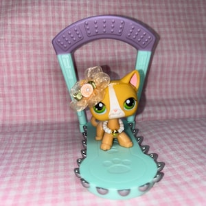 May include: A small orange and white cat toy with a light green paw print on the base. The cat is wearing a light orange and yellow flower headband and a gold necklace. The toy is sitting on a light purple and light green plastic swing set.