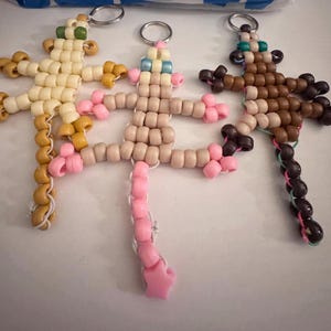Könnte beinhalten: Drei Schlüsselanhänger in Form von Eidechsen aus Perlen in den Farbtönen Beige, Rosa und Braun. Jede Eidechse hat einen Metall-Schlüsselring. Die Schlüsselanhänger sind handgefertigt und weisen eine Vielzahl von Perlenfarben und -mustern auf.
