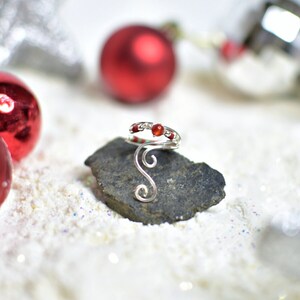 May include: A silver ring with a red stone and white beads. The ring is on a gray rock with a white background.