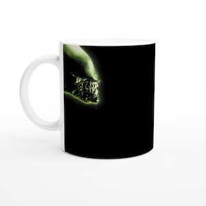 May include: White ceramic mug with a black and green alien creature graphic. The alien's mouth is open and its teeth are visible. The background is black.