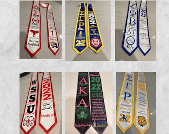 Puede incluir: Seis estolas de graduación con diferentes colores, diseños y textos. Las estolas están hechas de satén y tienen varios colores, incluyendo rojo, blanco, azul, negro, amarillo y verde. Las estolas presentan diferentes textos, incluyendo letras griegas, nombres de escuelas y el año de graduación. Las estolas se muestran sobre un fondo blanco.