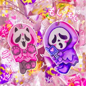 May include: Two colorful cartoon character charms with a pink and purple color scheme. The charms are of a ghost holding a phone and a panda wearing a pink hooded robe. The charms are surrounded by pink and white candy wrappers with Japanese text.