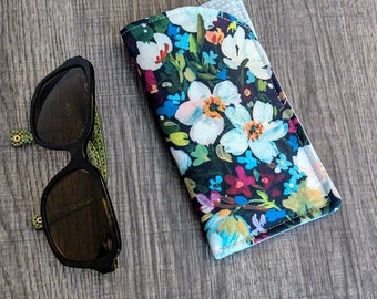 May include: A fabric spectacles case featuring a dark background with a vibrant floral print in shades of white, light blue, pink, and green, positioned next to a pair of black-framed sunglasses with green patterned arms.