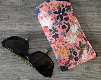 May include: A rectangular fabric spectacle case featuring a pink background with a multi-coloured floral pattern of white, yellow, blue, and green flowers, paired with black-framed sunglasses that have green patterned arms and dark lenses.