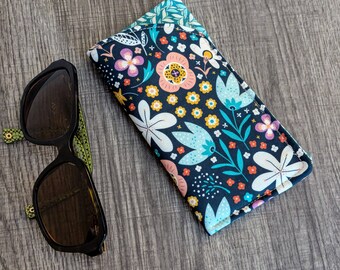 May include: A rectangular fabric spectacle case featuring a dark navy blue background with a colourful floral pattern of white, light blue, pink, and yellow flowers and leaves, positioned next to a pair of black sunglasses with green patterned arms.