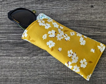 May include: A rectangular fabric spectacles case featuring a mustard yellow background with a white floral pattern. The case is partially open, revealing a pair of black-framed sunglasses tucked inside with a glimpse of a green patterned lining.