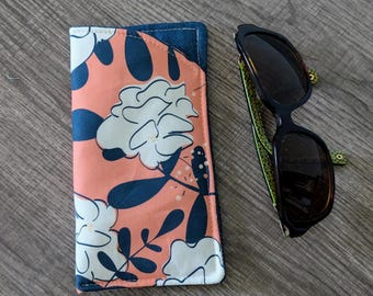 May include: A rectangular fabric spectacle case featuring a peach-coloured background with a dark blue and white floral pattern, positioned next to a pair of black-framed sunglasses with green patterned inner arms and dark tinted lenses.