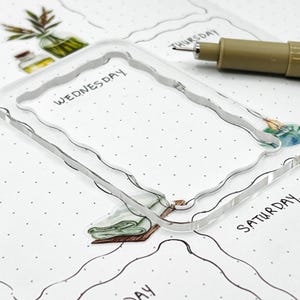 May include: A clear acrylic stamp with a wavy border and the word "WEDNESDAY" written on it. The stamp is on a white sheet of paper with a dotted pattern.