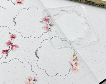 May include: A clear plastic template with a scalloped edge, used for creating decorative shapes in a planner or journal. The template is placed on a page with a dotted grid pattern, and there are several hand-drawn scalloped shapes on the page.