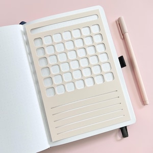 May include: Open notebook with a beige stencil overlay featuring a calendar grid and lined sections. A light pink pen rests beside the notebook on a pink surface. The notebook pages have a dotted grid pattern.