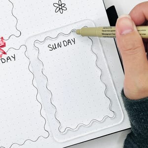 May include: A clear plastic stencil with a wavy border. The stencil is placed on a white dotted planner page. The word "SUNDAY" is written in black ink inside the stencil.