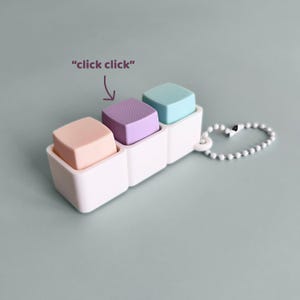 Puede incluir: Un juguete de plástico blanco con tres compartimentos cuadrados, cada uno con un cubo de diferente color: melocotón, lavanda y verde azulado. El juguete tiene una cadena de cuentas y el texto "click click" con una flecha que apunta al cubo central.