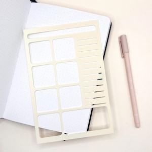 Peut inclure: Un pochoir crème avec des découpes rectangulaires et linéaires, placé sur une page de carnet à pois. Un stylo rose clair est posé à côté du pochoir et du carnet. Le stylo porte le numéro 8.