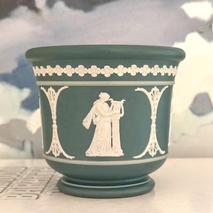 May include: A matte sage green ceramic planter featuring white relief ornamentation. The design includes a continuous floral border at the rim and vertical columns separating a central figure playing a lyre. The vessel has a flared rim and base.