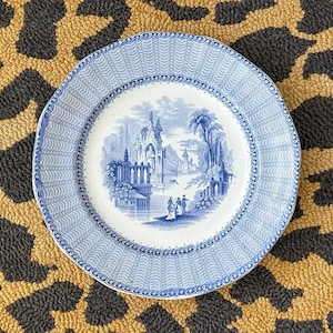 May include: A round ceramic plate featuring a blue and white transferware pattern. The center depicts a detailed landscape with figures, buildings, and a body of water. The wide rim has a repeating wave-like texture and a decorative border.