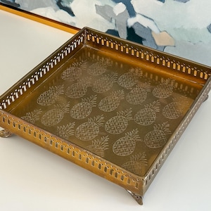 May include: A square, gold-colored metal tray with a decorative pineapple pattern etched into the base. The tray has a pierced border and rests on small, rounded feet. The tray is likely used for serving or display.