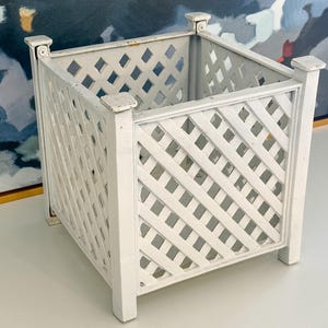 May include: A square, white, lattice-style planter box. The box has a classic design with a diamond pattern and is supported by four corner posts. The planter is made of wood and is painted white. The box is empty.