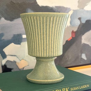 May include: A light green ceramic vase with a pedestal base and a flared, ribbed top. The vase is sitting on a dark green book. The background features an abstract painting with blue, gray, and white colors.