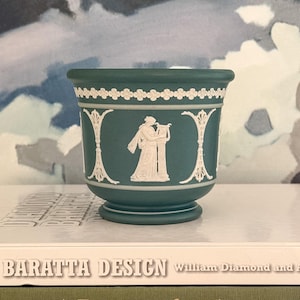 May include: A teal-colored ceramic planter with white relief detailing. The planter features a classical figure playing a harp, surrounded by decorative white accents. The planter is sitting on a white surface.