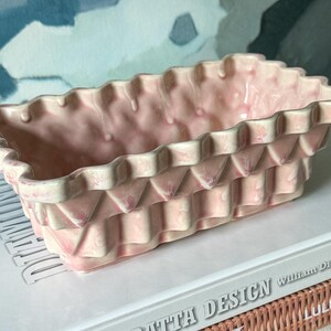 May include: A rectangular ceramic planter with a light pink and white marbled glaze. The exterior features a textured, basket-weave pattern with a scalloped, wavy top rim. The vessel has a deep, open interior and a flat base for stability.