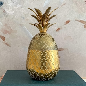 May include: A metallic gold pineapple-shaped container with a removable lid. The body features a textured, diamond-patterned crosshatch design, while the top is adorned with pointed, upright leaves. The object has a polished, reflective finish.