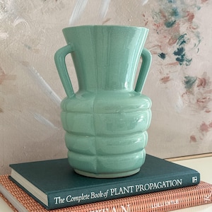 May include: A light mint green ceramic vase with a flared rim and two side handles. The body of the vase features a segmented, grid-like texture. The vase is positioned on top of a dark green book titled The Complete Book of PLANT PROPAGATION.