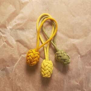 May include: Three decorative zipper pulls made of woven cord in shades of bright yellow, mustard yellow, and olive green. Each pull features a monkey fist knot at the end of a looped cord, creating a textured, spherical design for easy gripping.