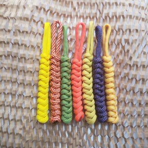 May include: Seven paracord zipper pulls arranged vertically, featuring braided designs in solid yellow, orange-and-white speckled, solid green, solid orange, solid light yellow, blue-and-white speckled, and solid golden yellow colors.
